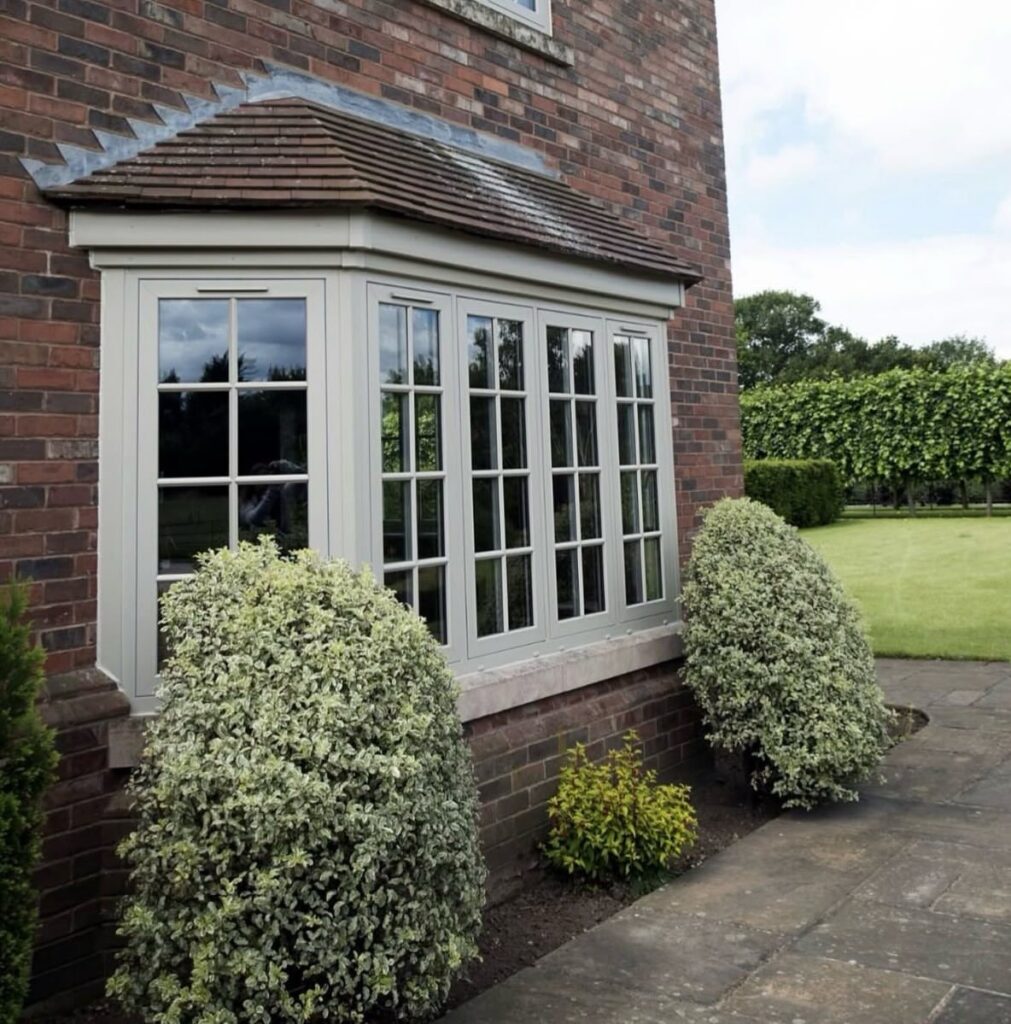 A grey bay window on a brick house surrounded by manicured green and white shrubs.