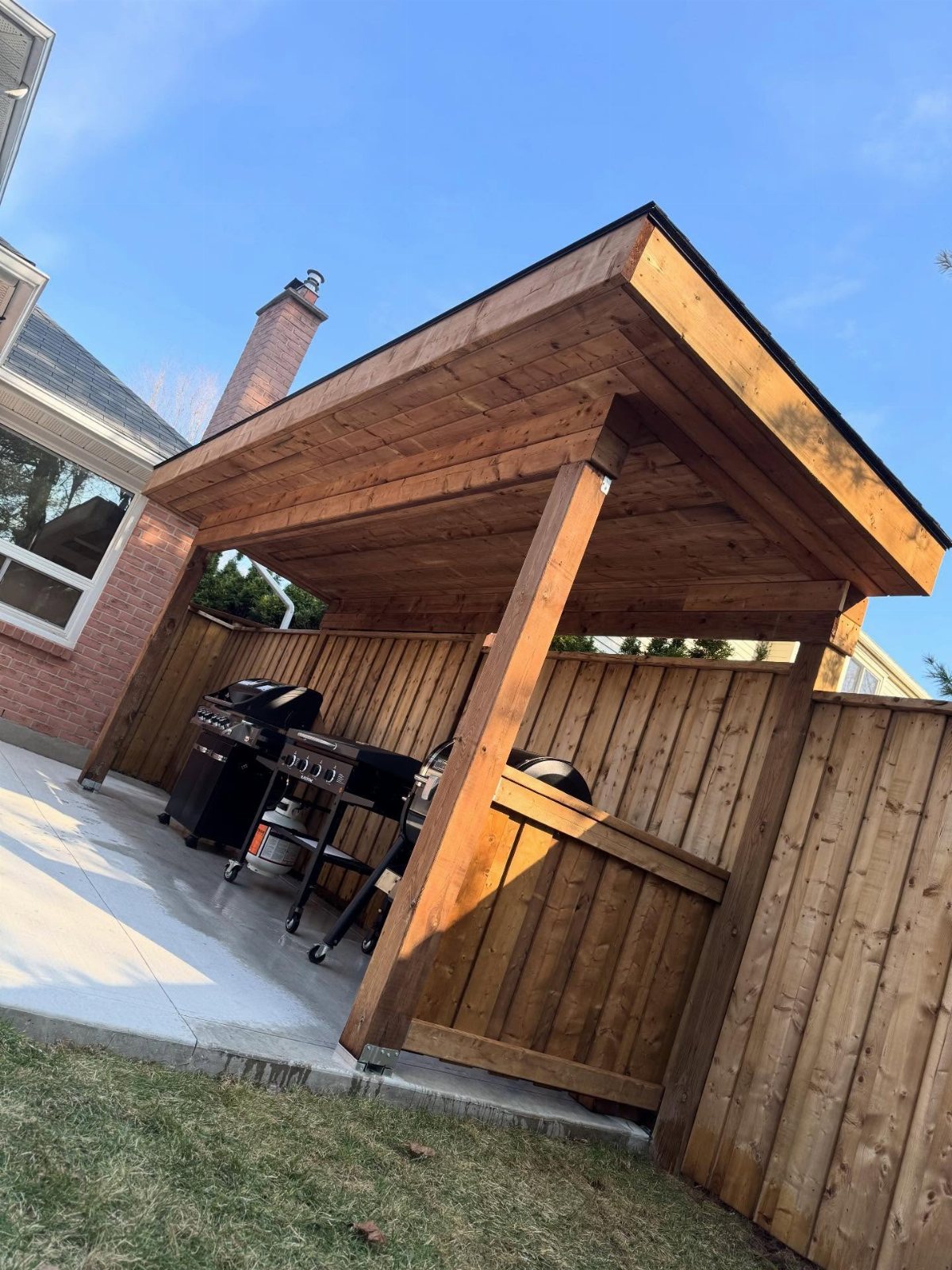 A wooden sheltered structure housing a black gas grill on a concrete patio.