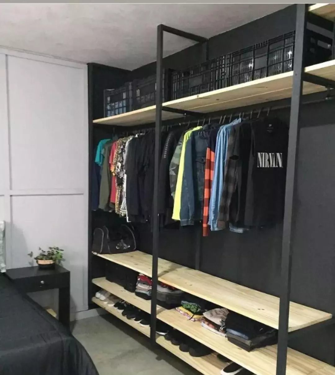 Black metal shelving unit with light wood shelves holding folded clothes, shoes, and hanging garments.