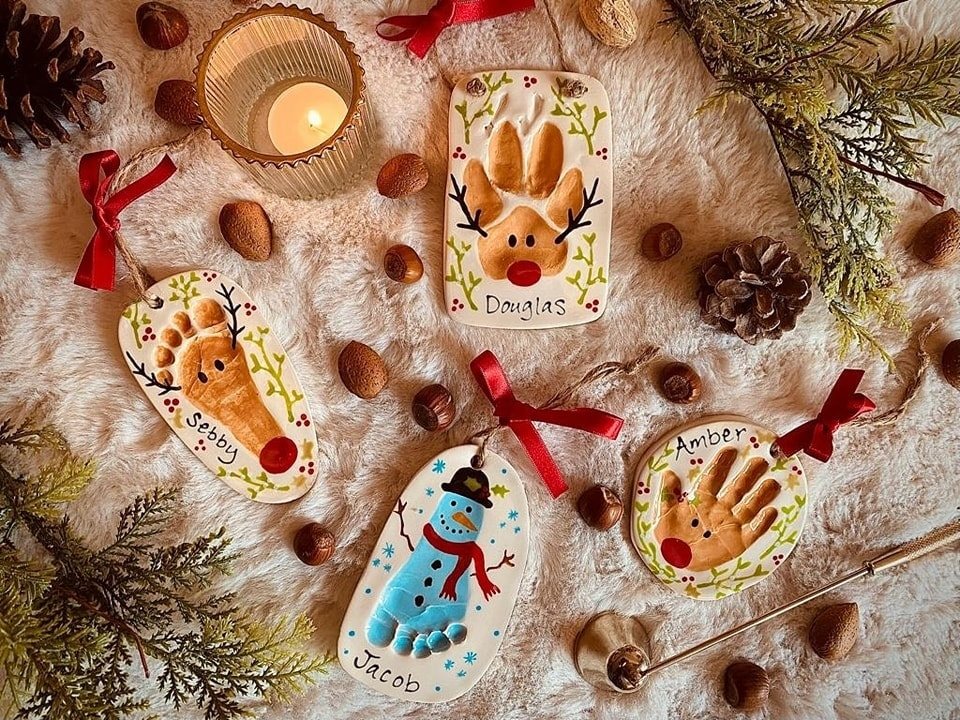 Four handmade clay ornaments with colorful fingerprint designs and names resting on white faux fur.