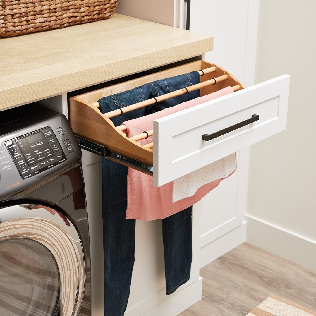A wooden pull-out drying rack extended from a white cabinet holding blue jeans and pink fabric.