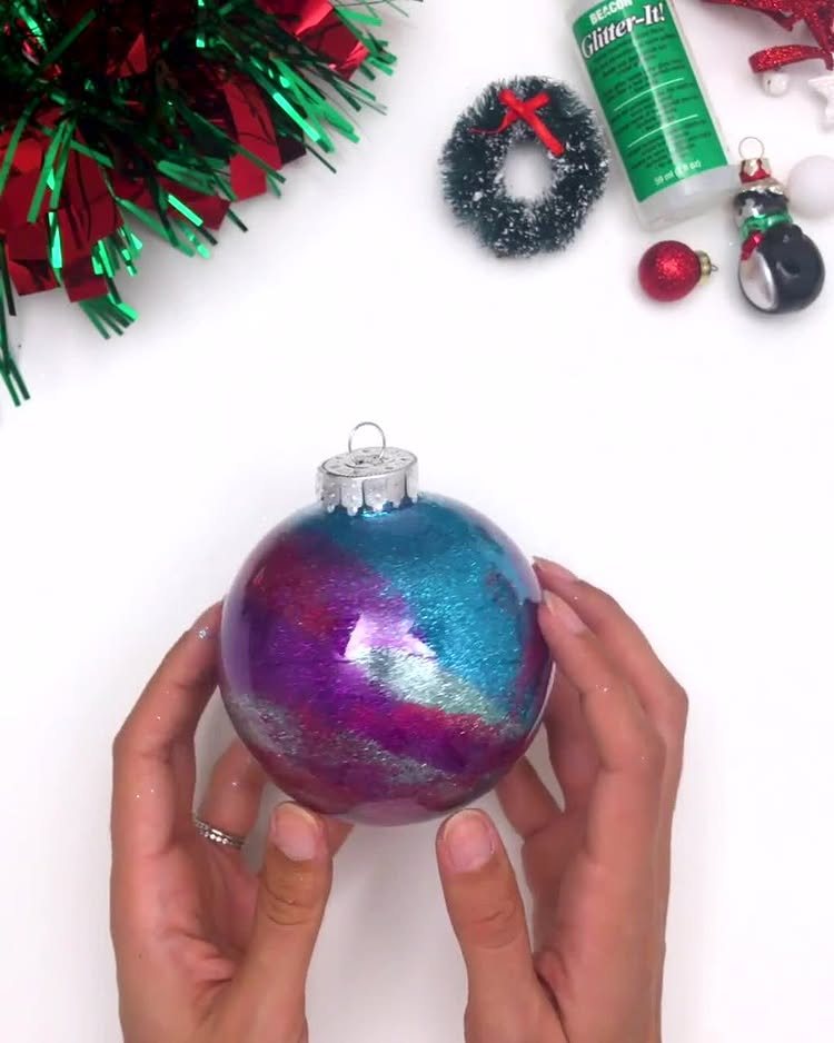 Hands holding a large Christmas ornament decorated with vibrant purple and blue glitter on a white background.