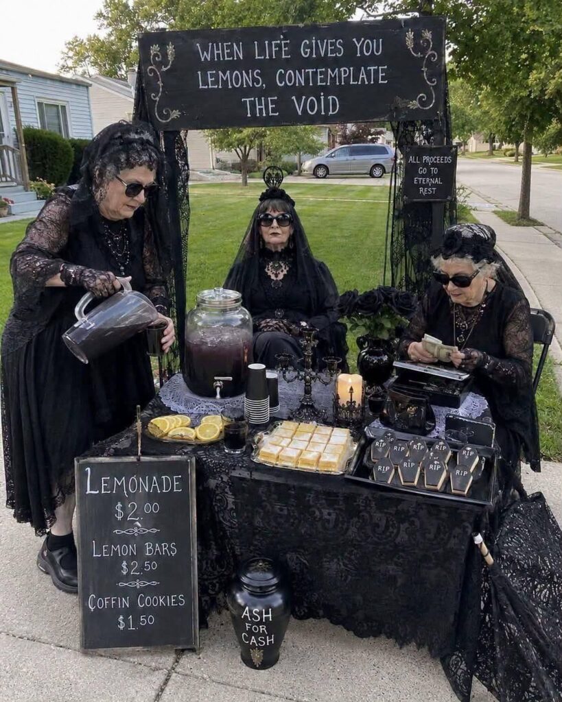 Three women in black lace clothing operating a gothic-themed lemonade stand outdoors.