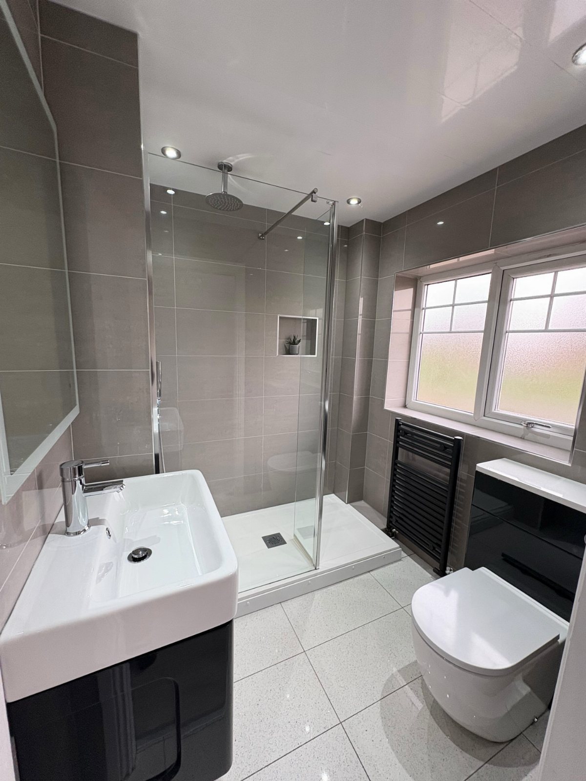 A modern bathroom with grey tiles, a glass shower, white sink, and toilet.
