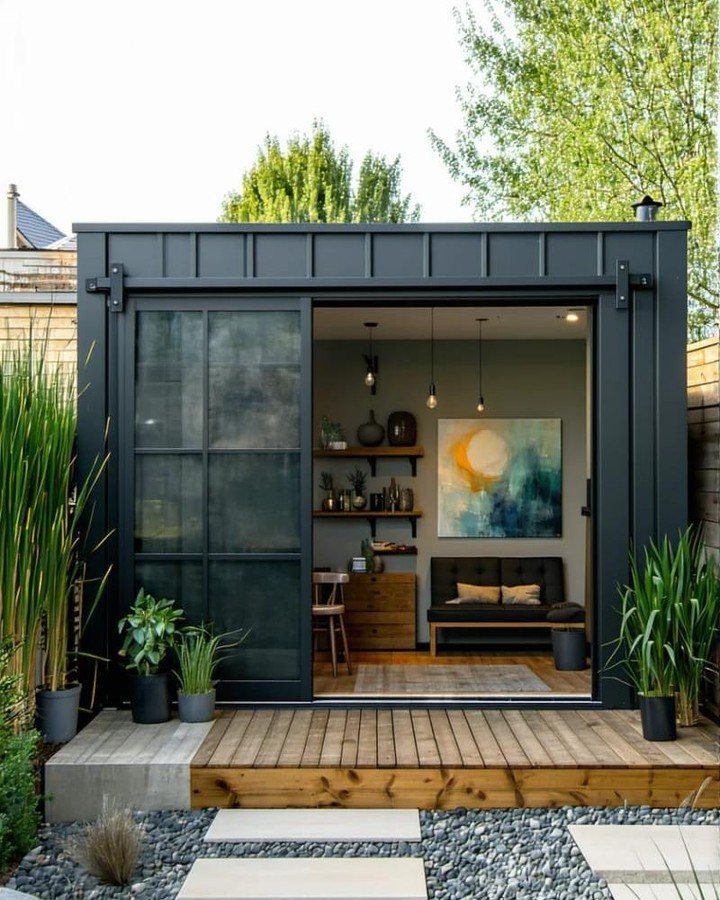 A modern dark grey backyard shed with glass doors opening to a furnished interior lounge.