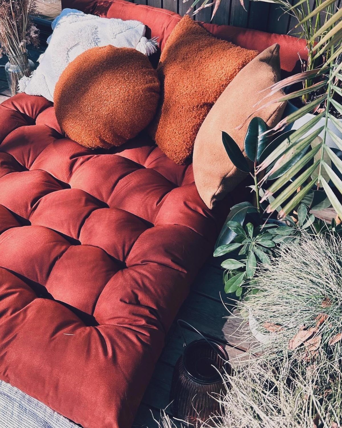 Reddish-orange tufted floor cushion with various orange and tan pillows on a sunny wooden deck.