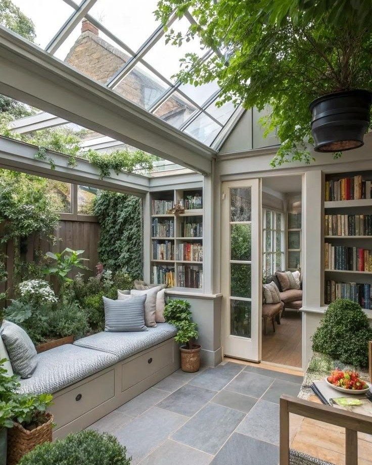 A bright sunroom with a glass ceiling, built-in bookshelves, and many green indoor plants.