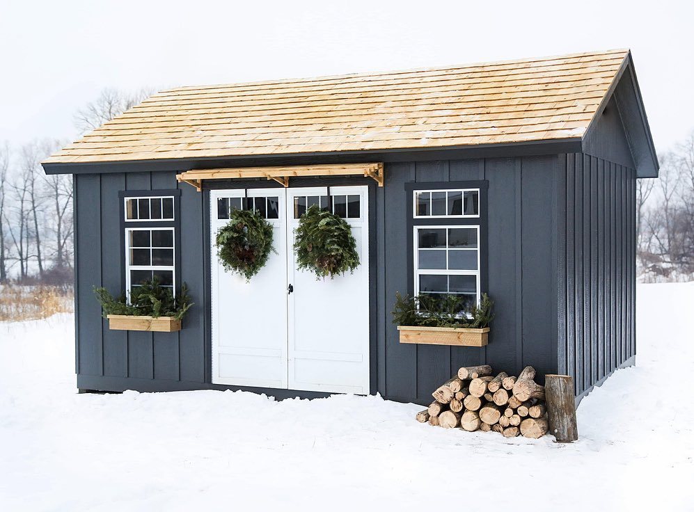 Charcoal gray wooden shed with white doors and windows sitting in a snowy field.