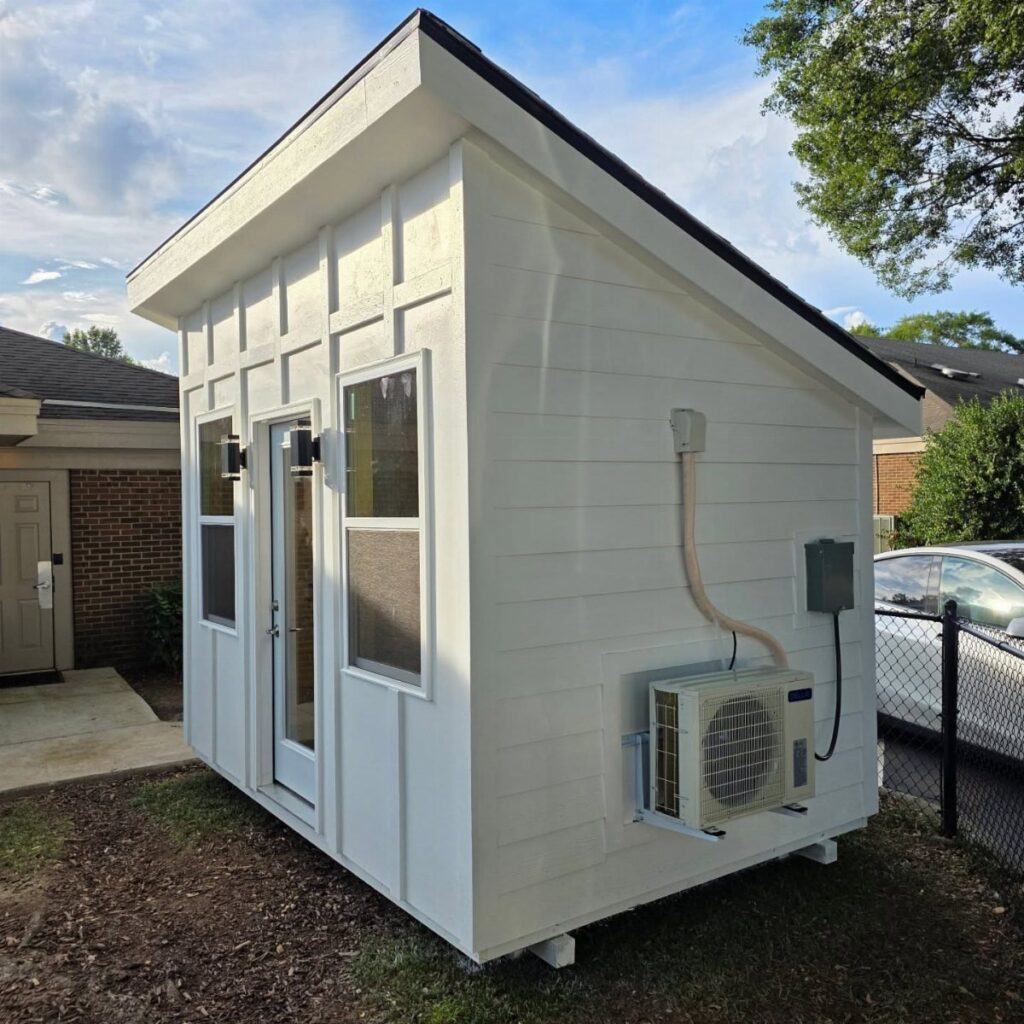 White wooden studio shed with an external air conditioning unit mounted on the side wall.