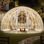A glowing transparent geodesic dome with string lights and outdoor furniture on a lawn at night.