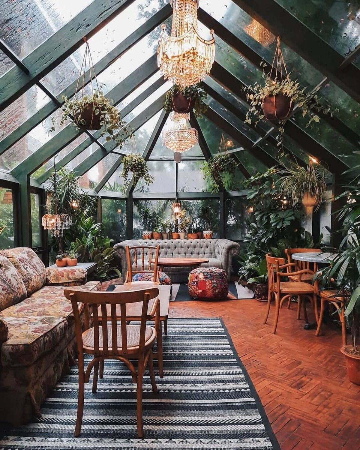 A lush conservatory room with glass walls, hanging plants, chandeliers, and comfortable seating areas.