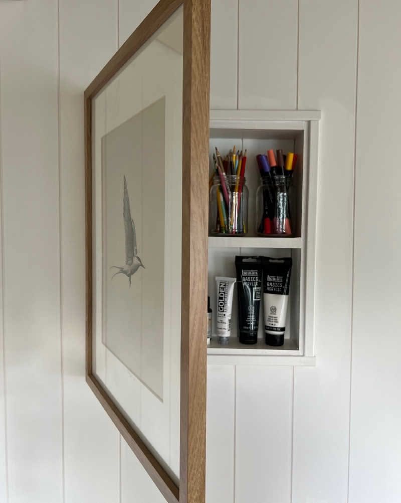 A wooden picture frame opened to reveal a white shadow box filled with art supplies.