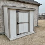 A white lean-to shed with tan trim attached to a brick building wall.