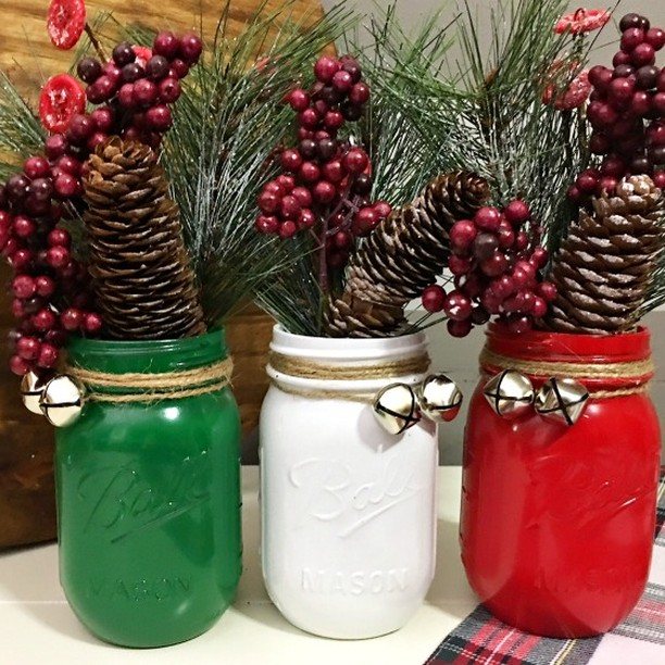 Three painted mason jars in green, white, and red holding festive pine and berry arrangements.