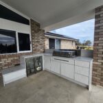 Modern outdoor kitchen with white cabinets, a built-in grill, and a small beverage fridge.